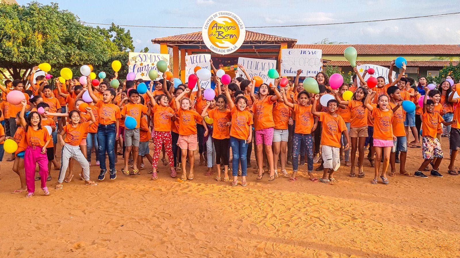 Alunos de camiseta laranja dos Amigos do Bem comemoram com balões e cartazes em frente ao Centro de Transformação da unidade de Inajá.